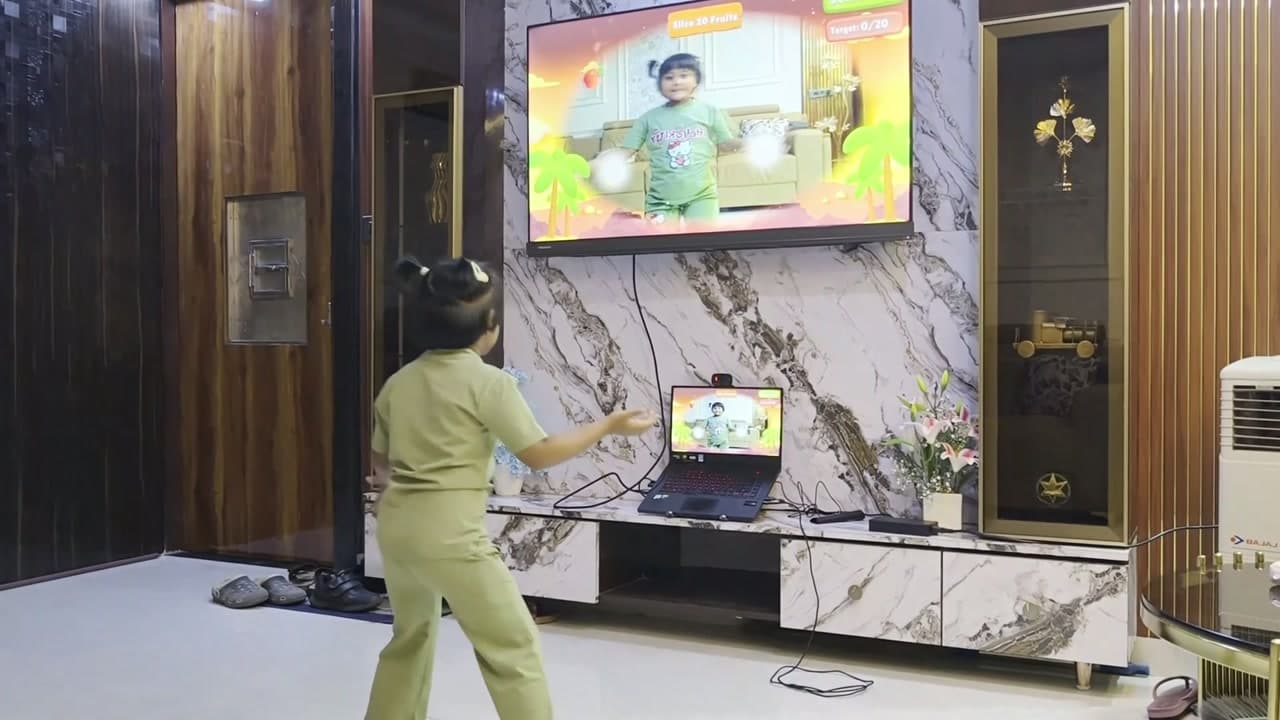 A laptop with its webcam on a TV stand, plugged into the TV, with a girl playing in front of it