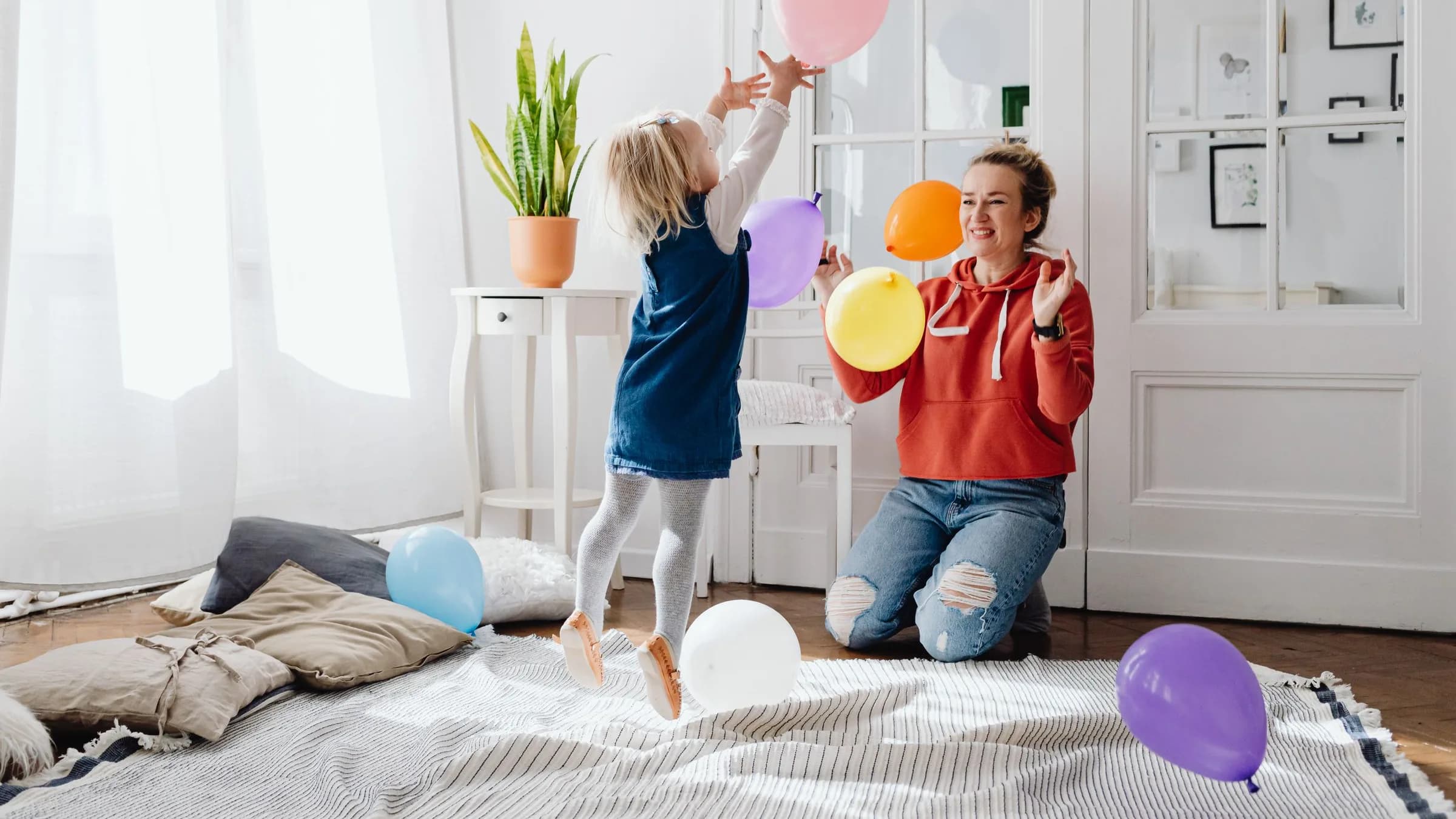 A preschool girl jumps for a pink balloon on a living-room rug while her mother kneels nearby, laughing