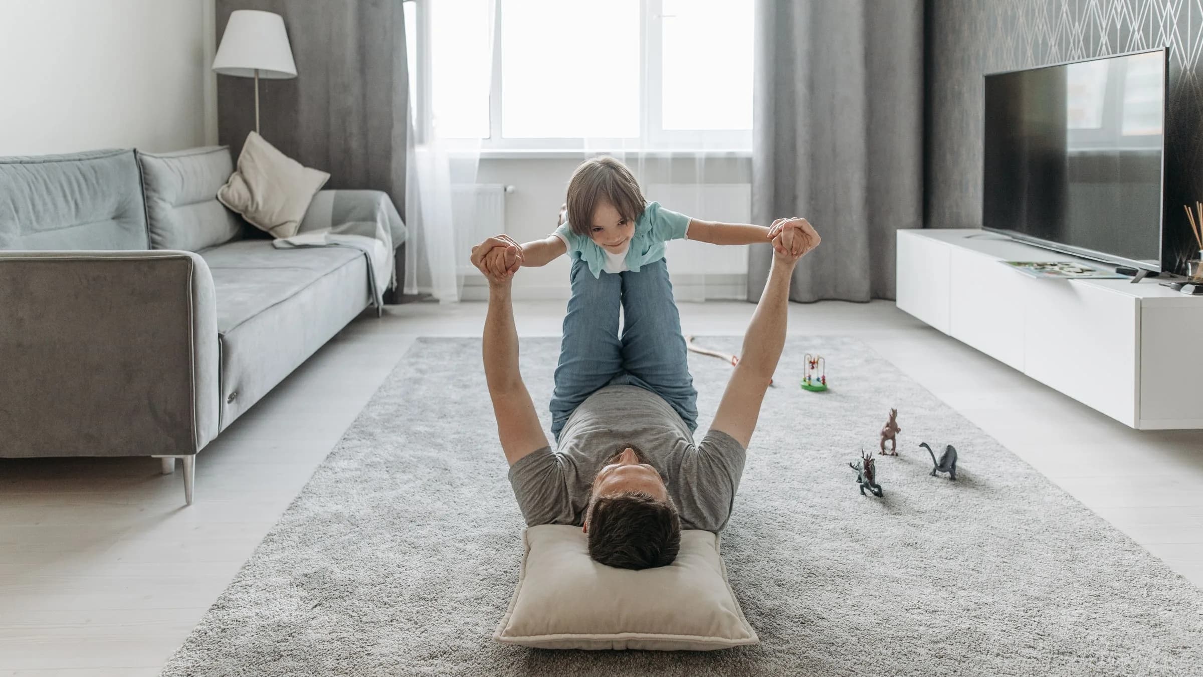 A father lies on a living-room rug and balances his preschool son in the air, toy dinosaurs scattered nearby