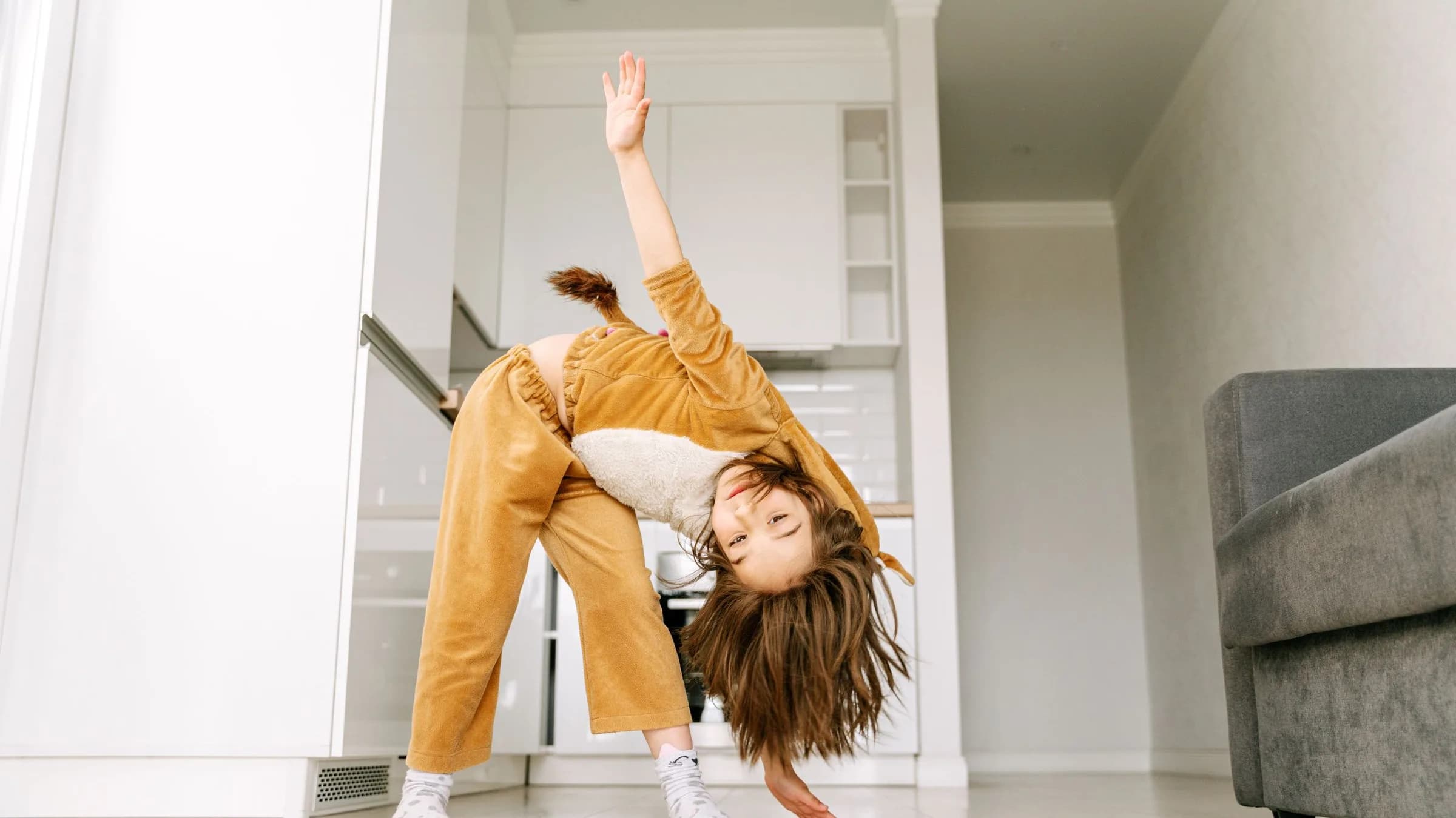 A girl of about 6 in lion-style pajamas bends to touch the floor with one hand, her other arm high, in a bright apartment