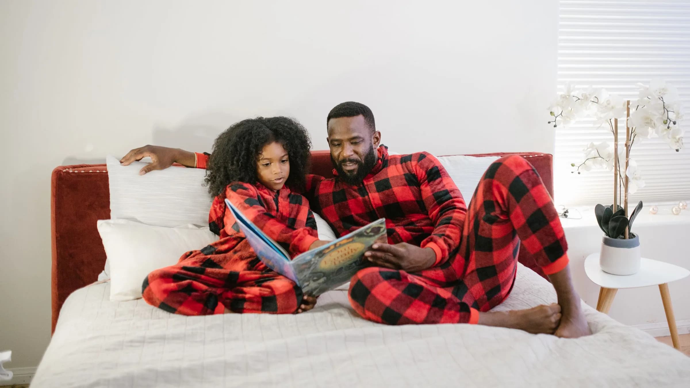 A father and his young daughter in matching plaid pajamas read a picture book together on a bed