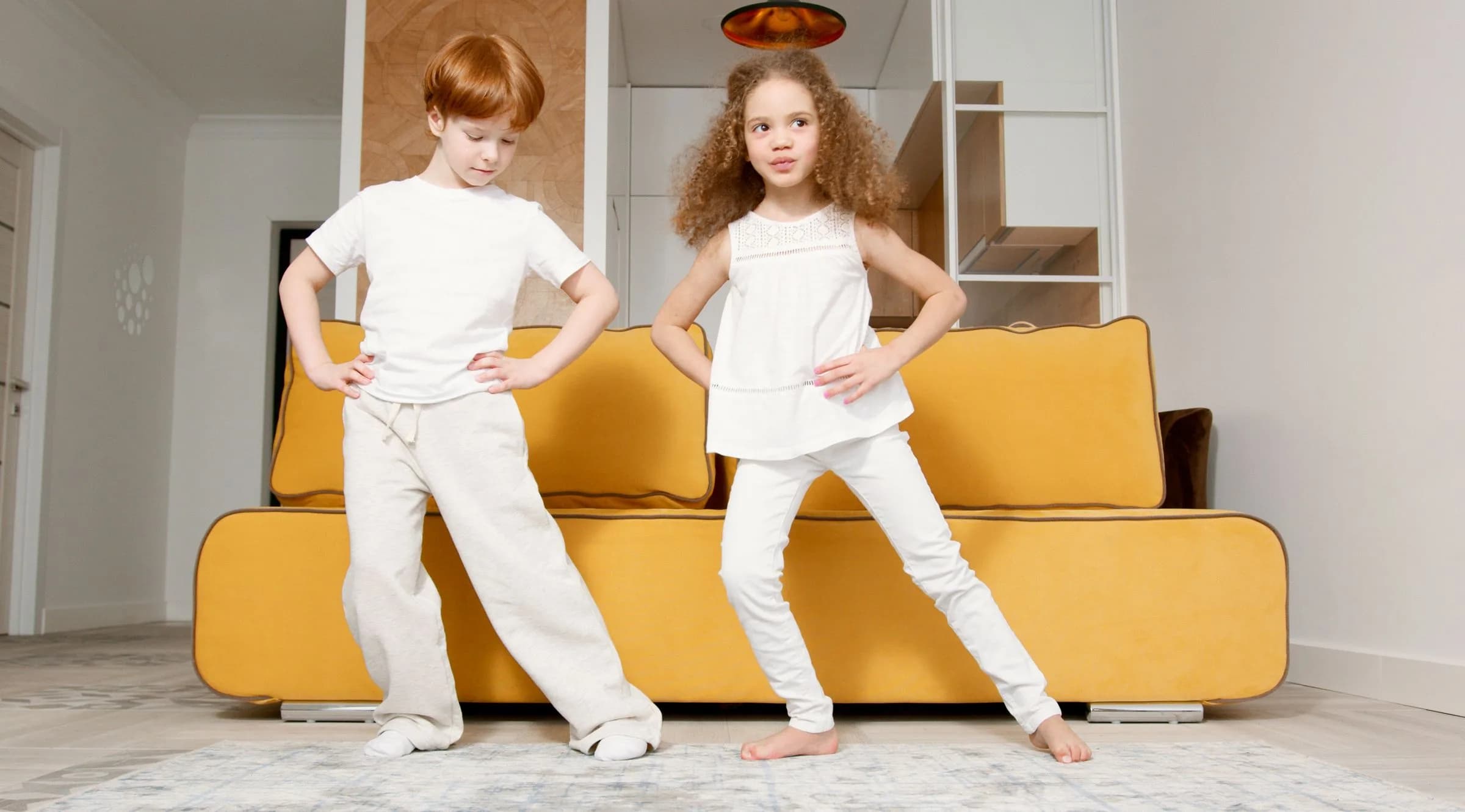 A boy and a girl of about 6, hands on hips, strike the same dance pose on a rug in front of a yellow sofa
